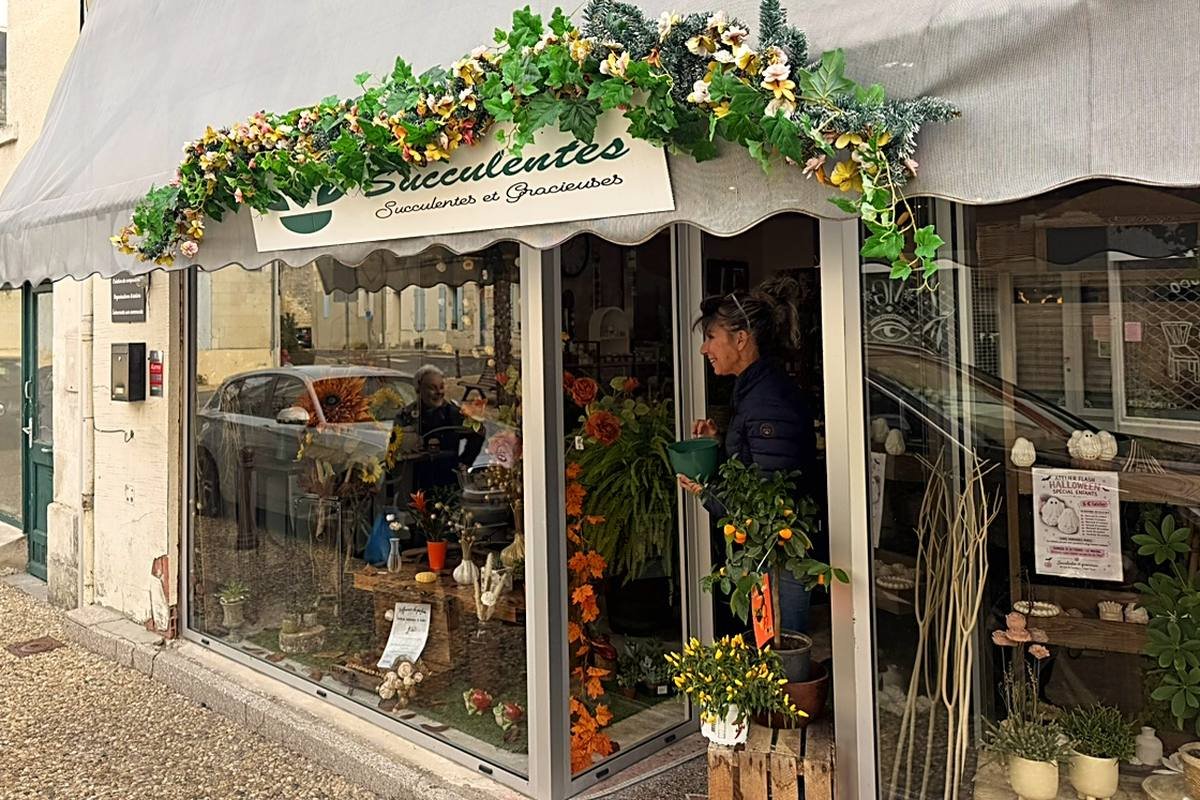 Devanture de la boutique Succulentes et gracieuses : enseigne sous l’auvent gris habillé d’une guirlande de fleurs et de lierre, vitrine garnie de fleurs et de plantes, porte ouverte sur la rue
