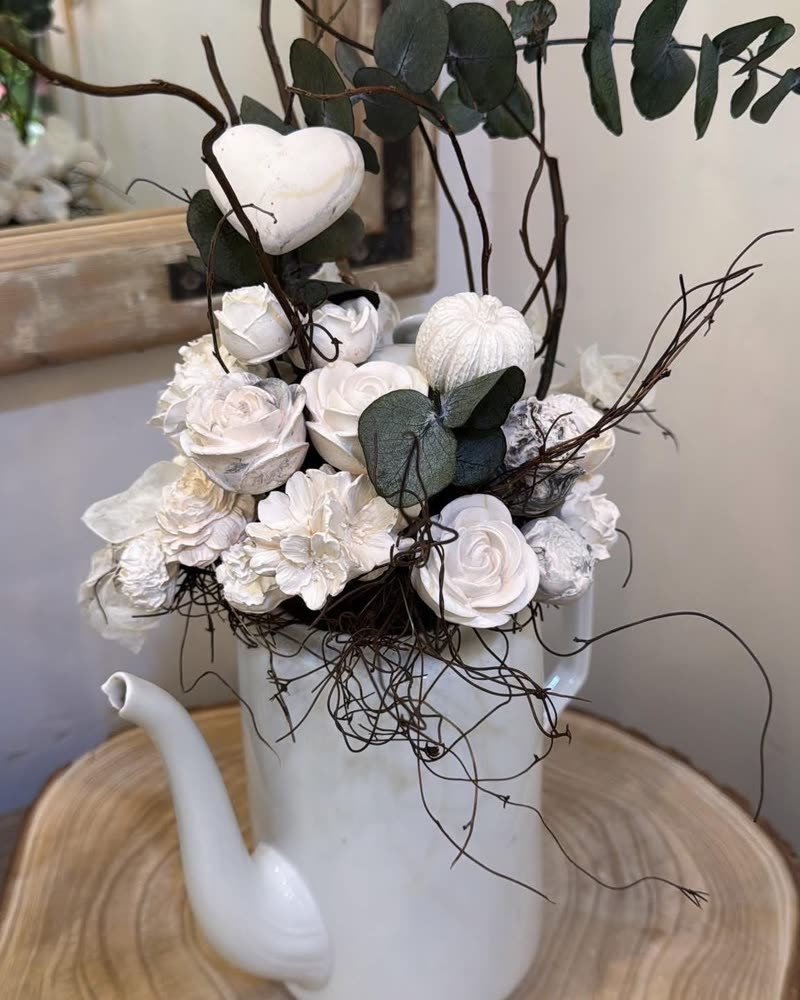 Ancienne cafetière en porcelaine blanche garnie de roses en gypse, d'eucalyptus et de branches de noisetier, posée sur une rondelle de bois